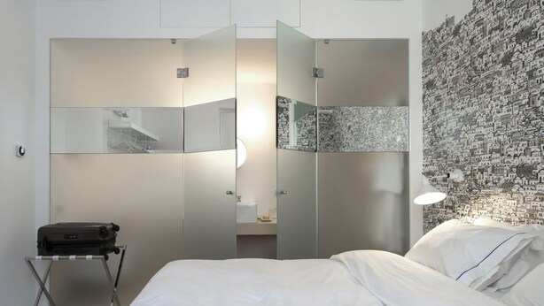 A hotel room with a bed, a suitcase on a stand, a mirror, and a wall with a cityscape wallpaper.