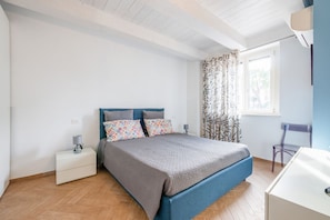 A bedroom with a bed, a nightstand, a chair, a window with curtains, and a desk.