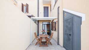 Un petit patio extérieur avec une table et des chaises en bois, un espace salon abrité et une porte grise.