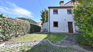Ein zweistöckiges Haus mit weißer Fassade, einer Holztür und einem kleinen Vorgarten mit befestigtem Bereich und einer Hecke.