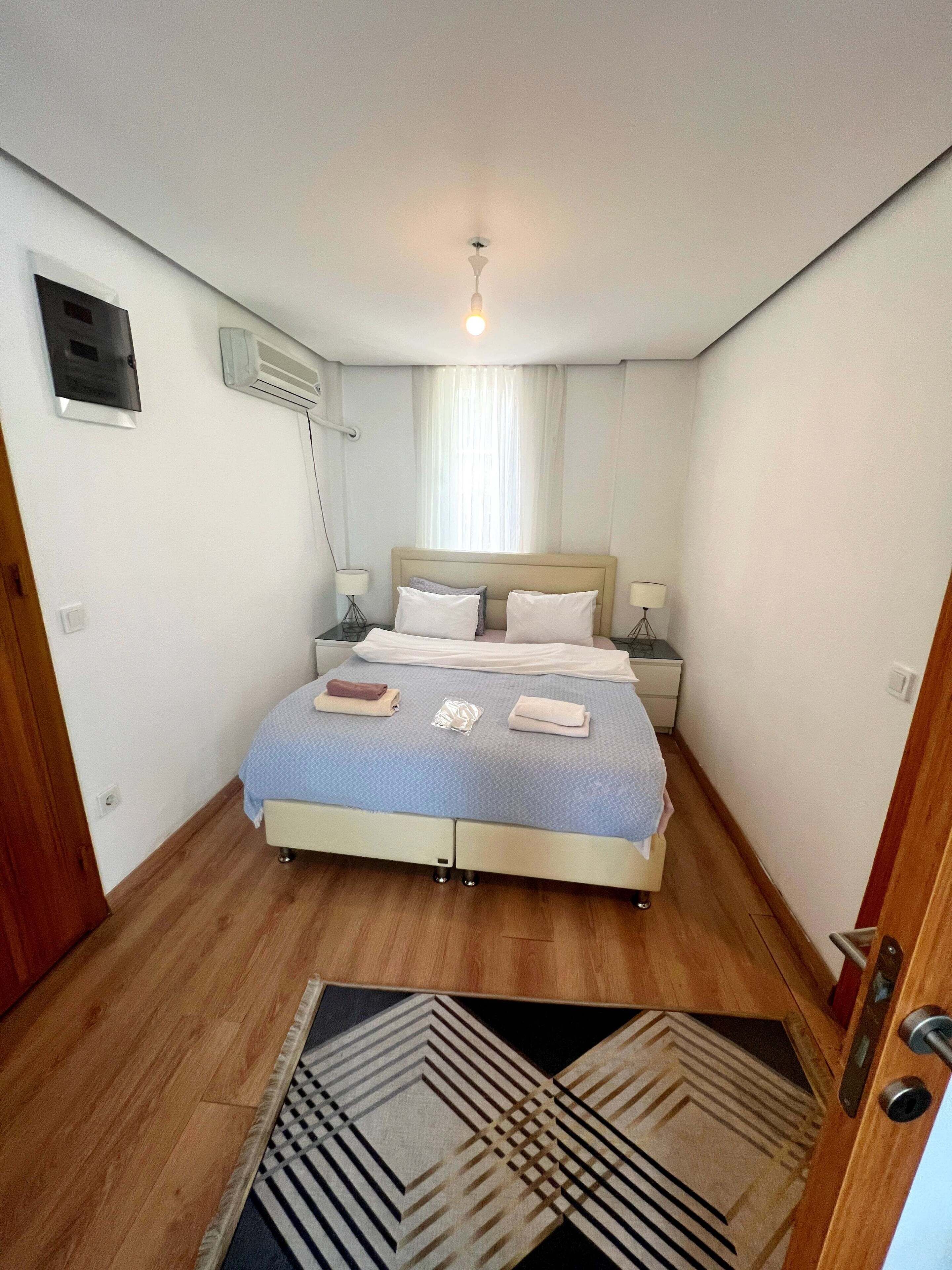 A bedroom with a bed, bedside tables, a ceiling light, an air conditioning unit, and a patterned rug.