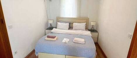 A bedroom with a bed, bedside tables, a ceiling light, an air conditioning unit, and a patterned rug.