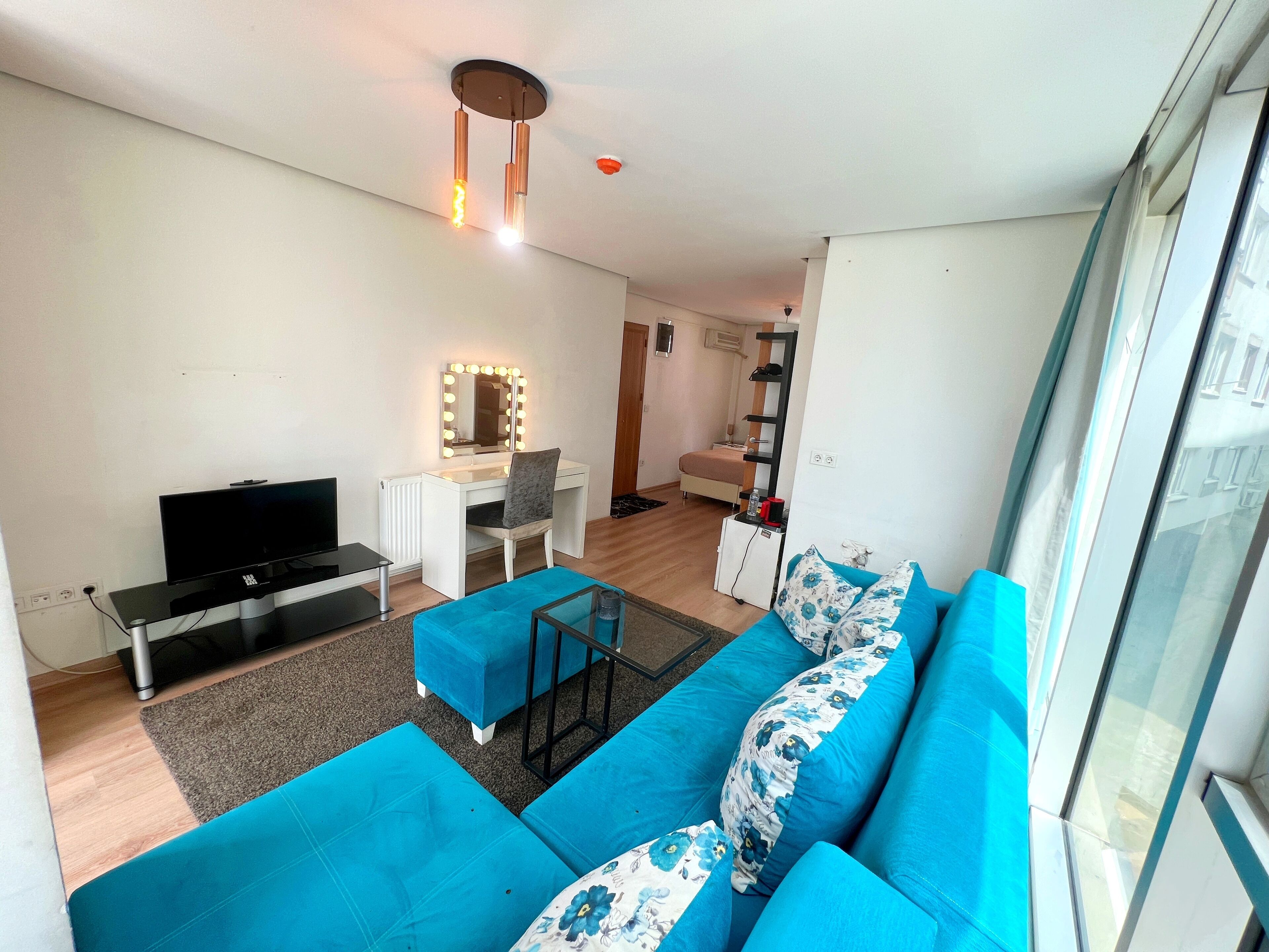 A modern living room with a sofa, a TV, a desk with a mirror, and a small kitchenette.