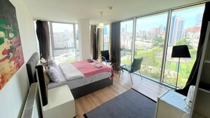 Ett modernt hotellrum med en stor säng, ett skrivbord och en stol. Genom de stora fönstren har man utsikt över staden.