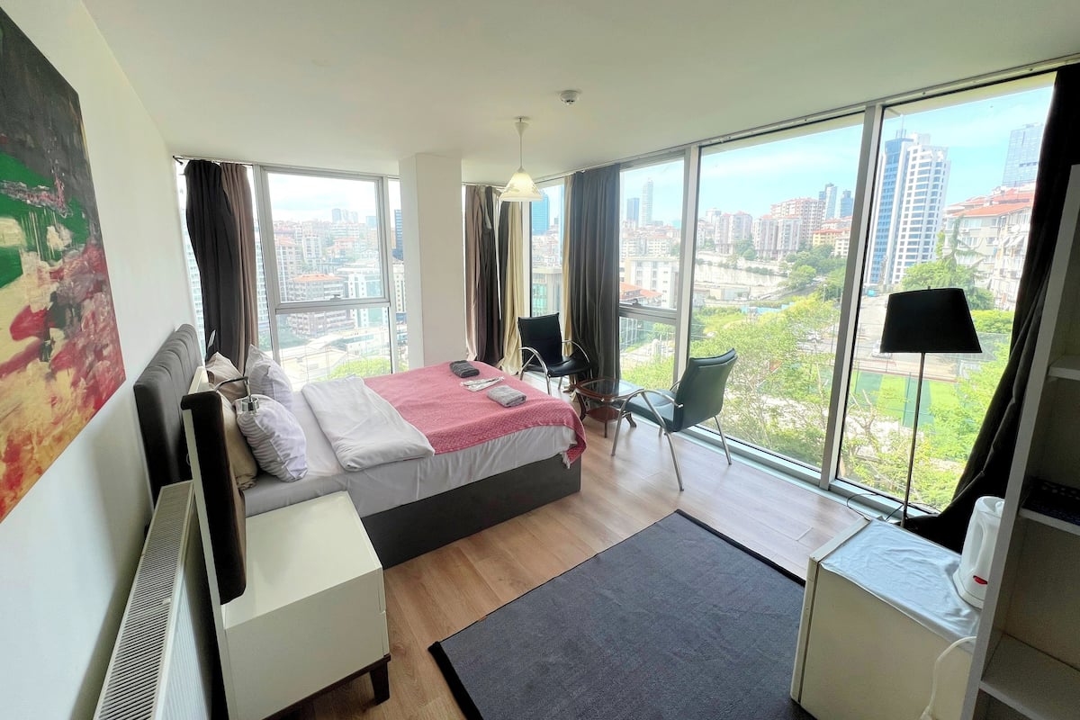 Quarto de hotel moderno com uma cama grande, uma escrivaninha e uma cadeira. Janelas amplas oferecem vista para a cidade.