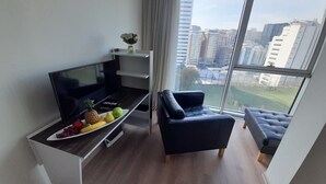 Ett modernt vardagsrum med en platt-TV, ett matbord med frukt, en soffa och ett stort fönster med utsikt över staden.
