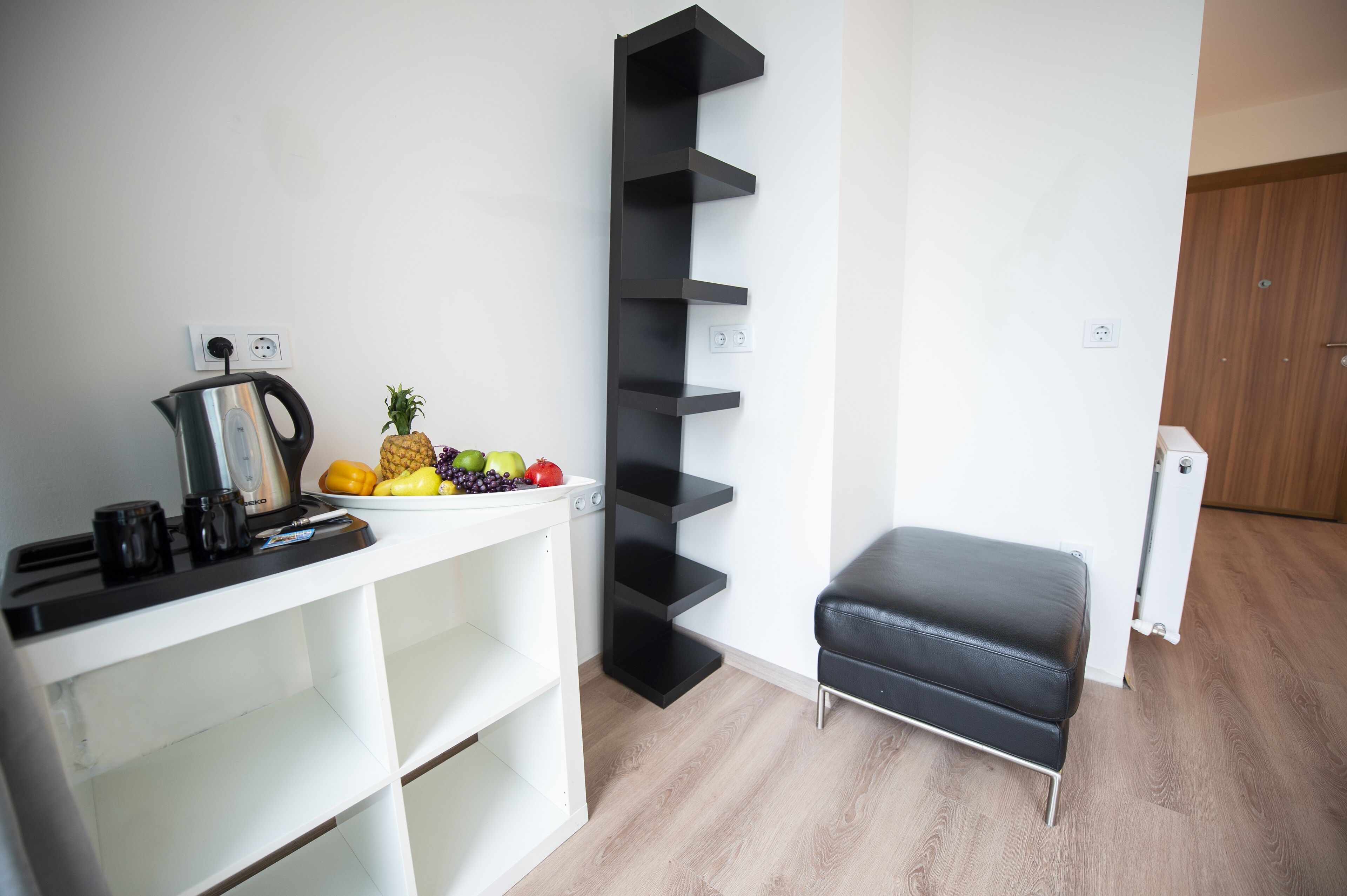 A kitchenette with a kettle, coffee maker, and fruit bowl. There is a black shelf unit and a black leather bench.