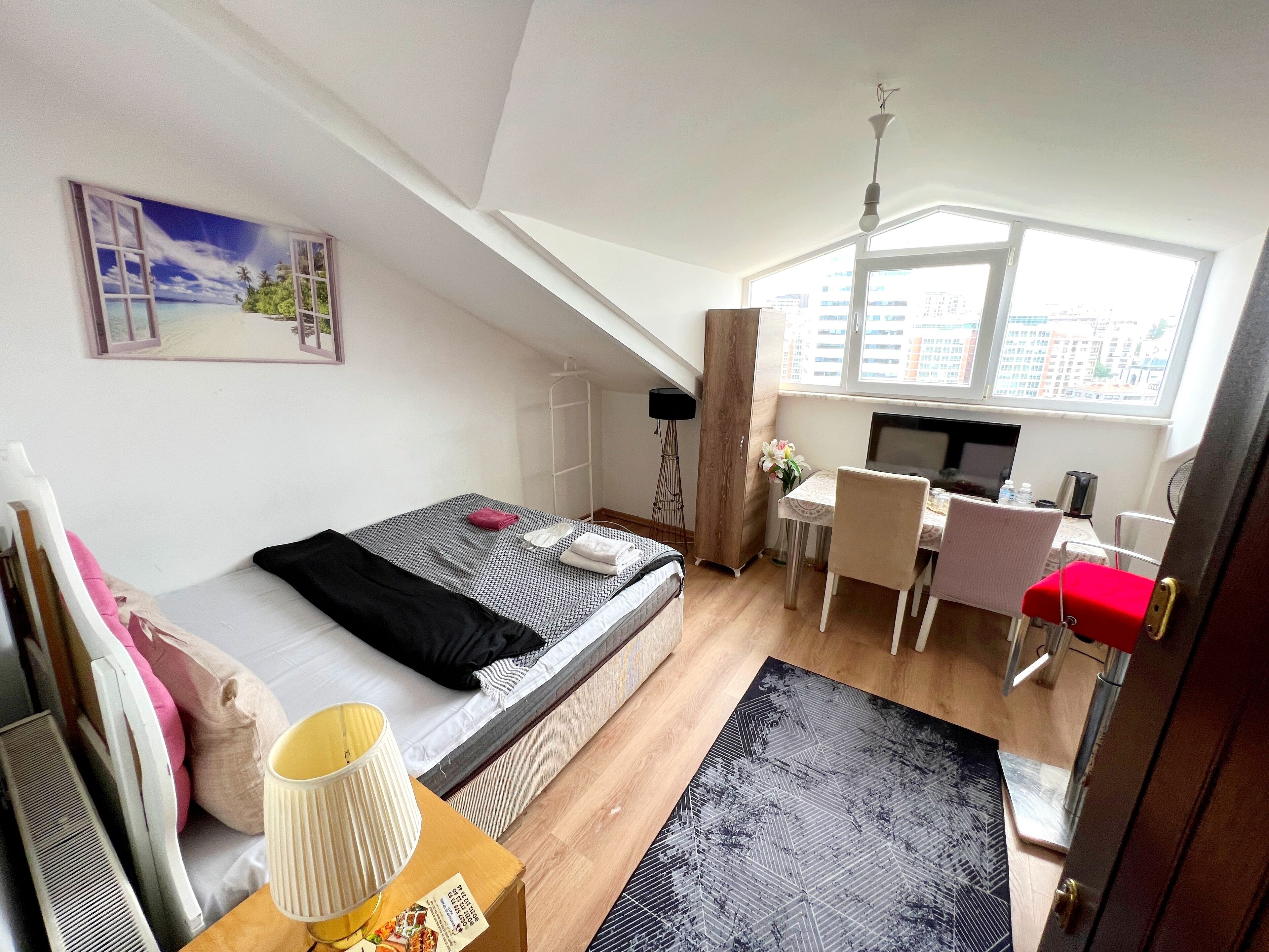 A compact room with a bed, a desk, and a chair. There is a window providing natural light and a view of buildings outside.