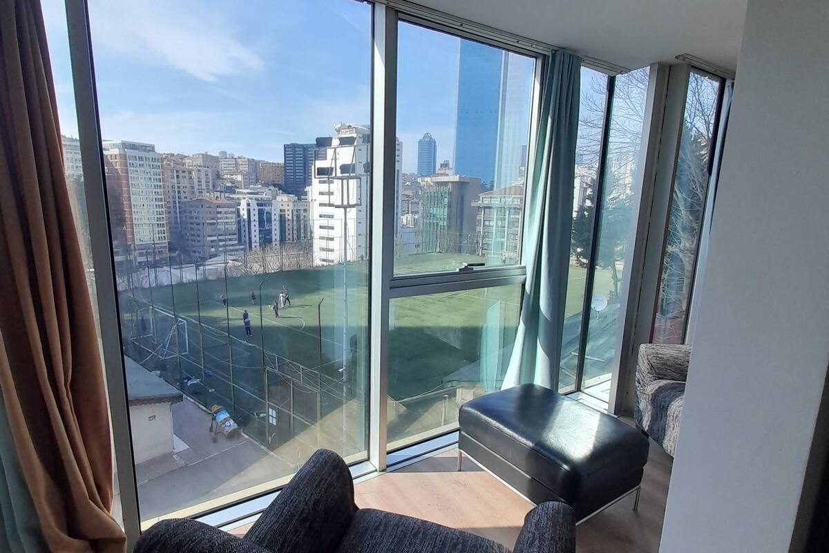Quarto moderno com amplas janelas do chão ao teto, sofá de couro preto e vista para a paisagem urbana, incluindo um campo esportivo.