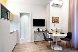 A modern kitchen with a built-in microwave, a dining area with a round table and two chairs, a flat-screen TV mounted on the wall, and a radiator under the window.