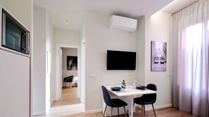 A modern dining area with a white table, two black chairs, a flat-screen TV mounted on the wall, and an air conditioning unit above.