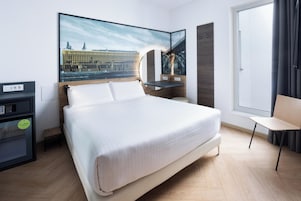 Una habitación de hotel moderna con una cama grande, un escritorio con computadora, una silla y un ventanal amplio.