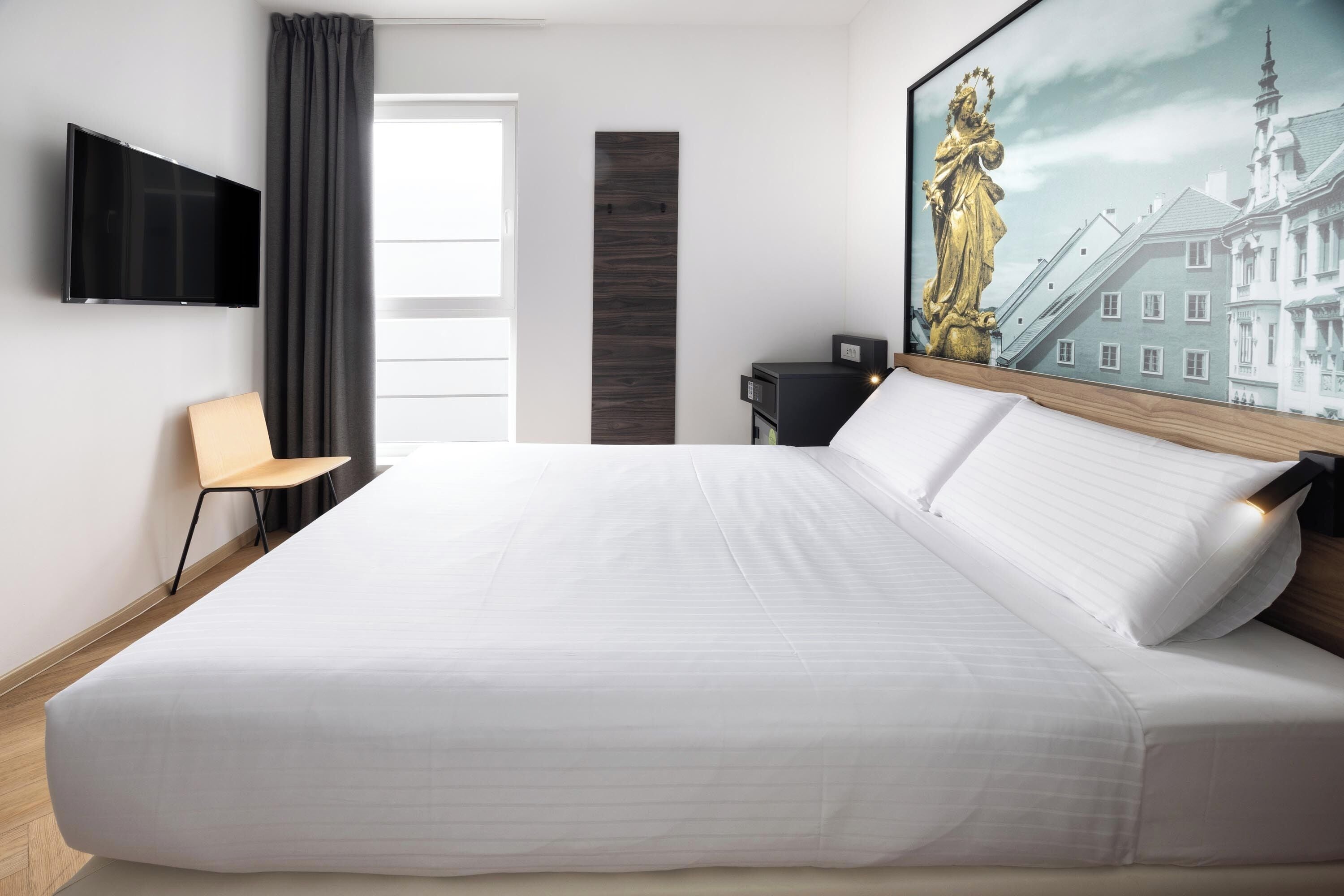 Une chambre d’hôtel moderne dotée d’un grand lit, d’un téléviseur à écran plat fixé au mur, d’un bureau avec une chaise et d’un tableau représentant un bâtiment.