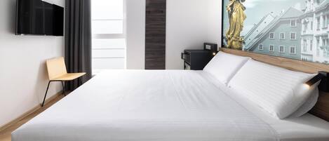 Une chambre d’hôtel moderne dotée d’un grand lit, d’un téléviseur à écran plat fixé au mur, d’un bureau avec une chaise et d’un tableau représentant un bâtiment.