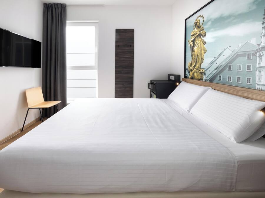 Ett modernt hotellrum med en stor säng, en platt-TV som är monterad på väggen, ett skrivbord med en stol och en tavla som föreställer en byggnad.