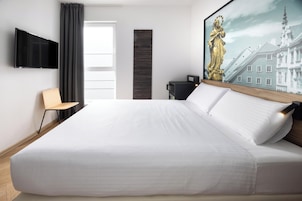 Una habitación de hotel moderna con una cama grande, un televisor de pantalla plana montado en la pared, un escritorio con una silla y un cuadro de un edificio.