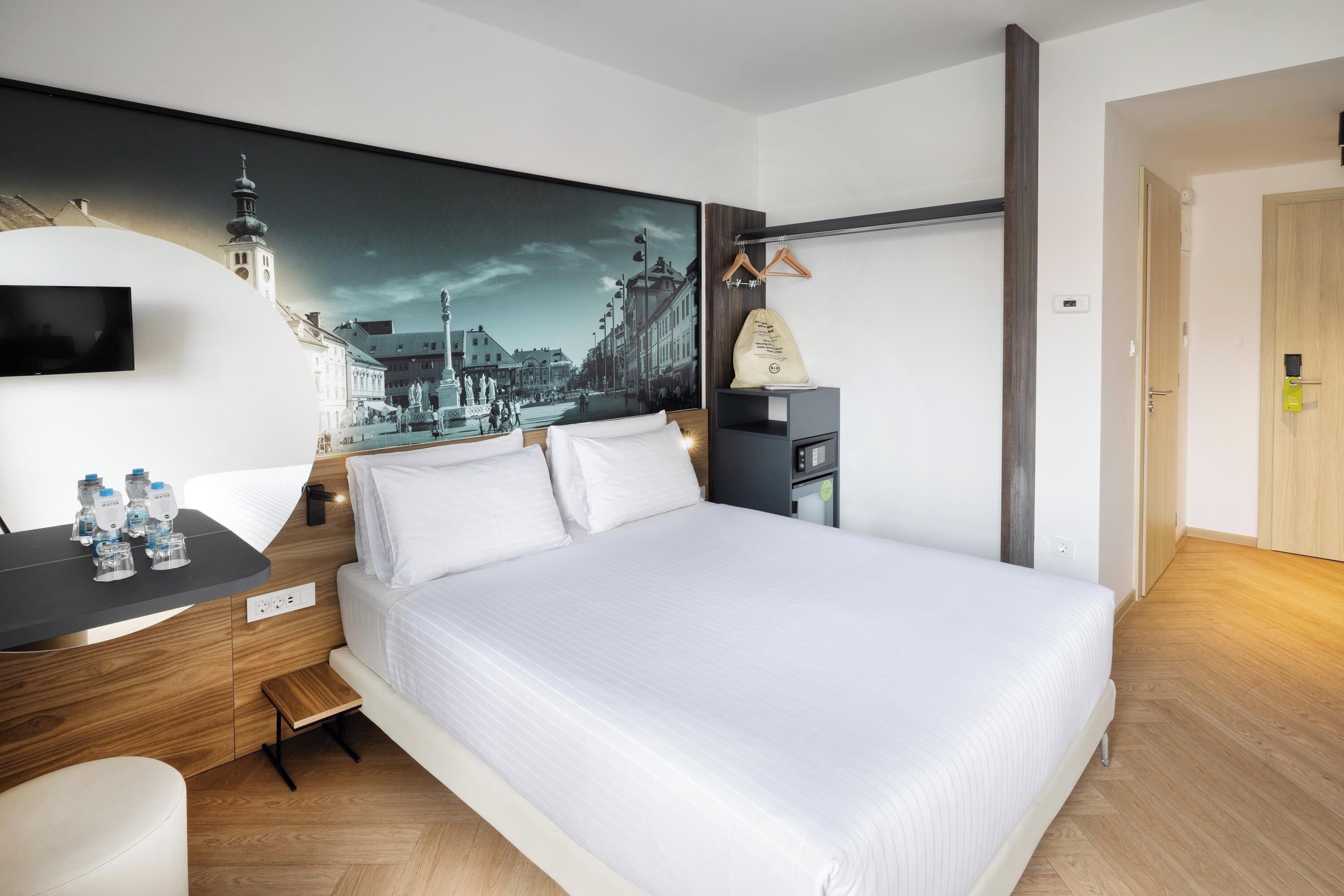 A hotel room with a bed, a desk with water bottles, a painting of a cityscape, and a coat hanging on a rack.