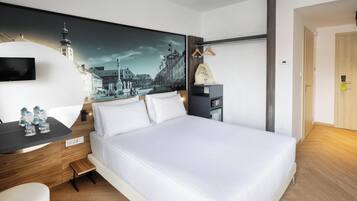 A hotel room with a bed, a desk with water bottles, a painting of a cityscape, and a coat hanging on a rack.