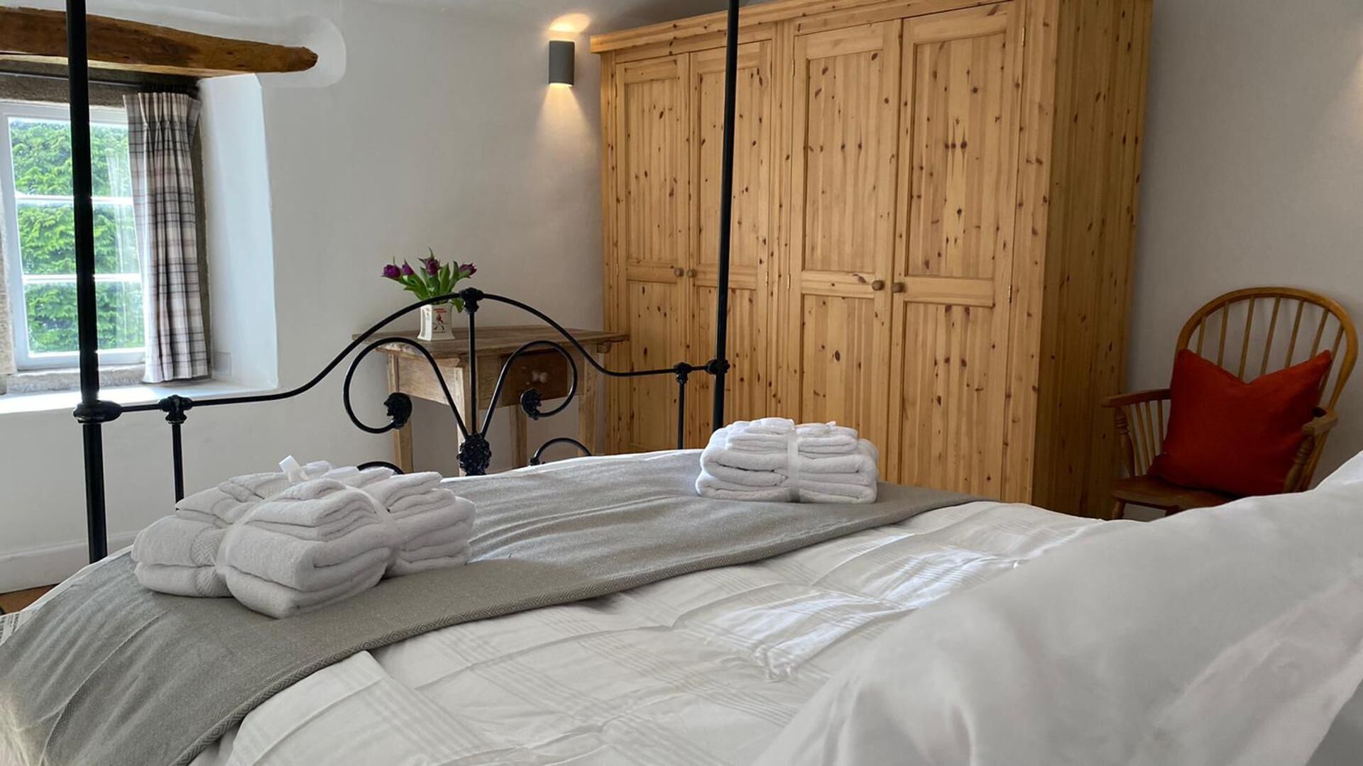 A neatly made bed with folded towels, a wooden wardrobe, a chair with a red cushion, and a window with curtains.