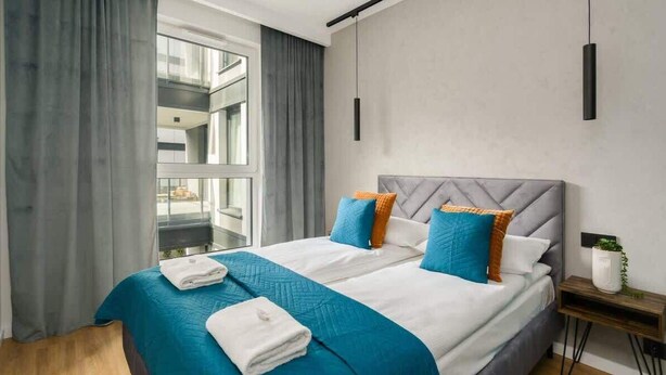 A modern hotel room with a large bed, grey curtains, a view of a balcony, and a small bedside table with a plant. - Cozy Apartment with Balcony | Parking | Close to Park (Poznan)