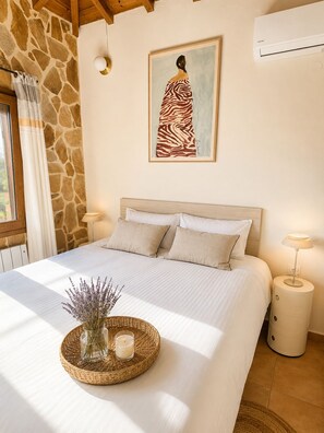 A bedroom with a stone wall, a bed with white bedding, a painting of a woman, and a small table with a candle and lavender. - Quiet villa with breathtaking views, garden and private pool! (Glossa)
