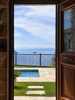A view through a door to a pool area with a clear sky, mountains, and a body of water. - Quiet villa with breathtaking views, garden and private pool! (Glossa)