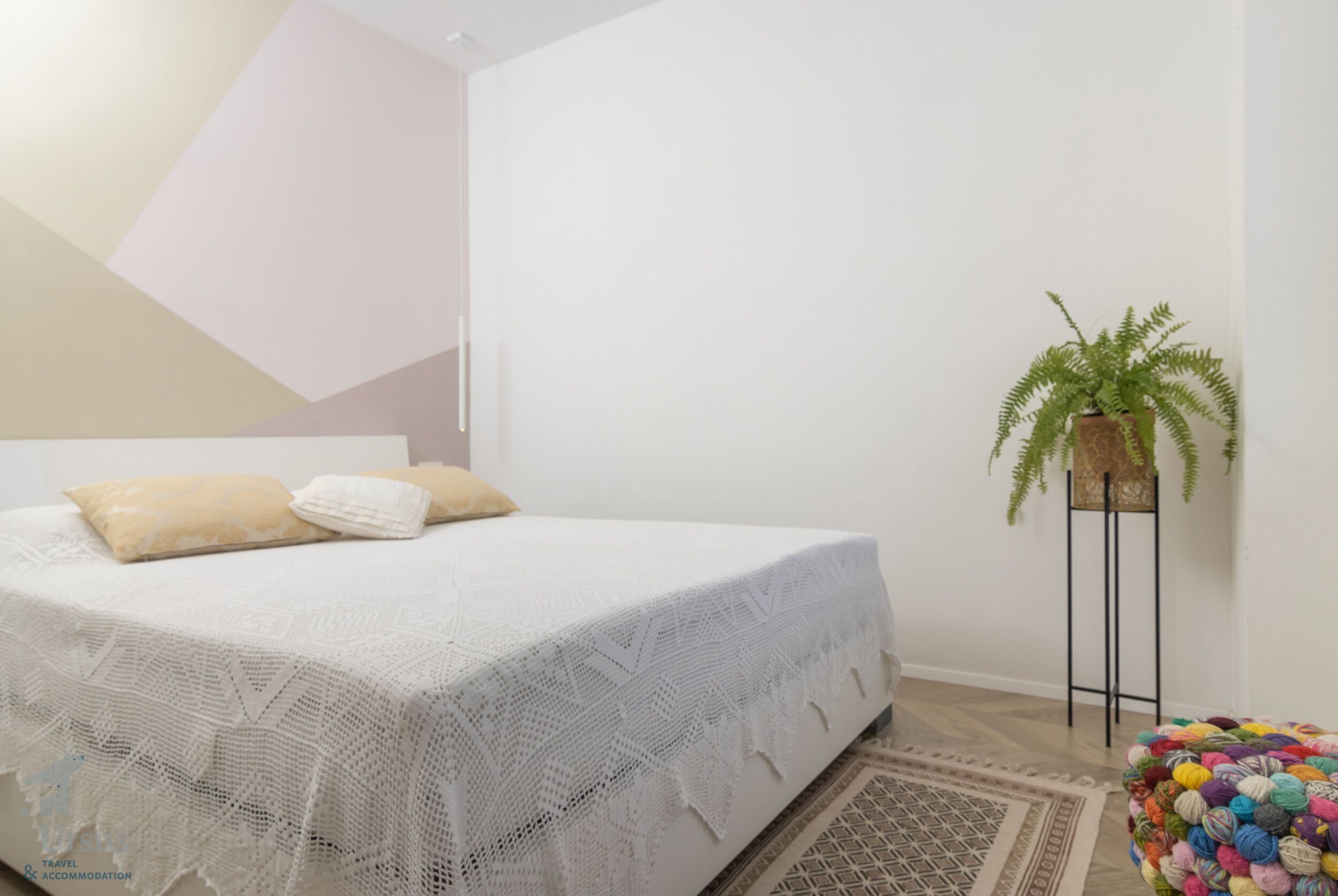 A neatly made bed with a white blanket and two pillows, a potted fern, and a colorful ball of yarn on a stand.