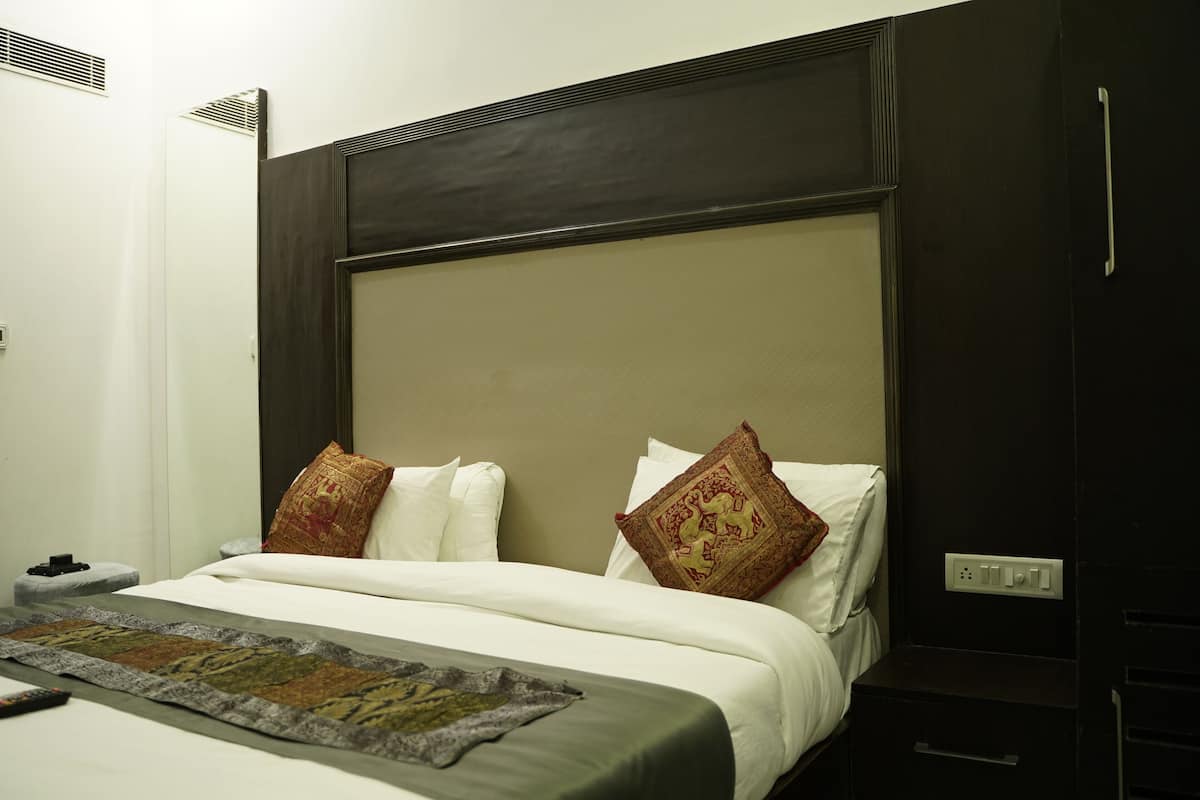Una cama bien hecha con sábanas blancas y almohadas decorativas, una mesita de noche con lámpara y un enchufe en la pared.