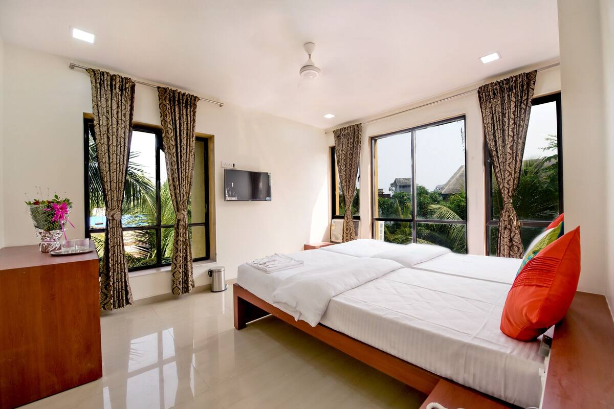 Quarto de hotel moderno com uma cama grande, uma escrivaninha, uma televisão e janelas amplas com cortinas.