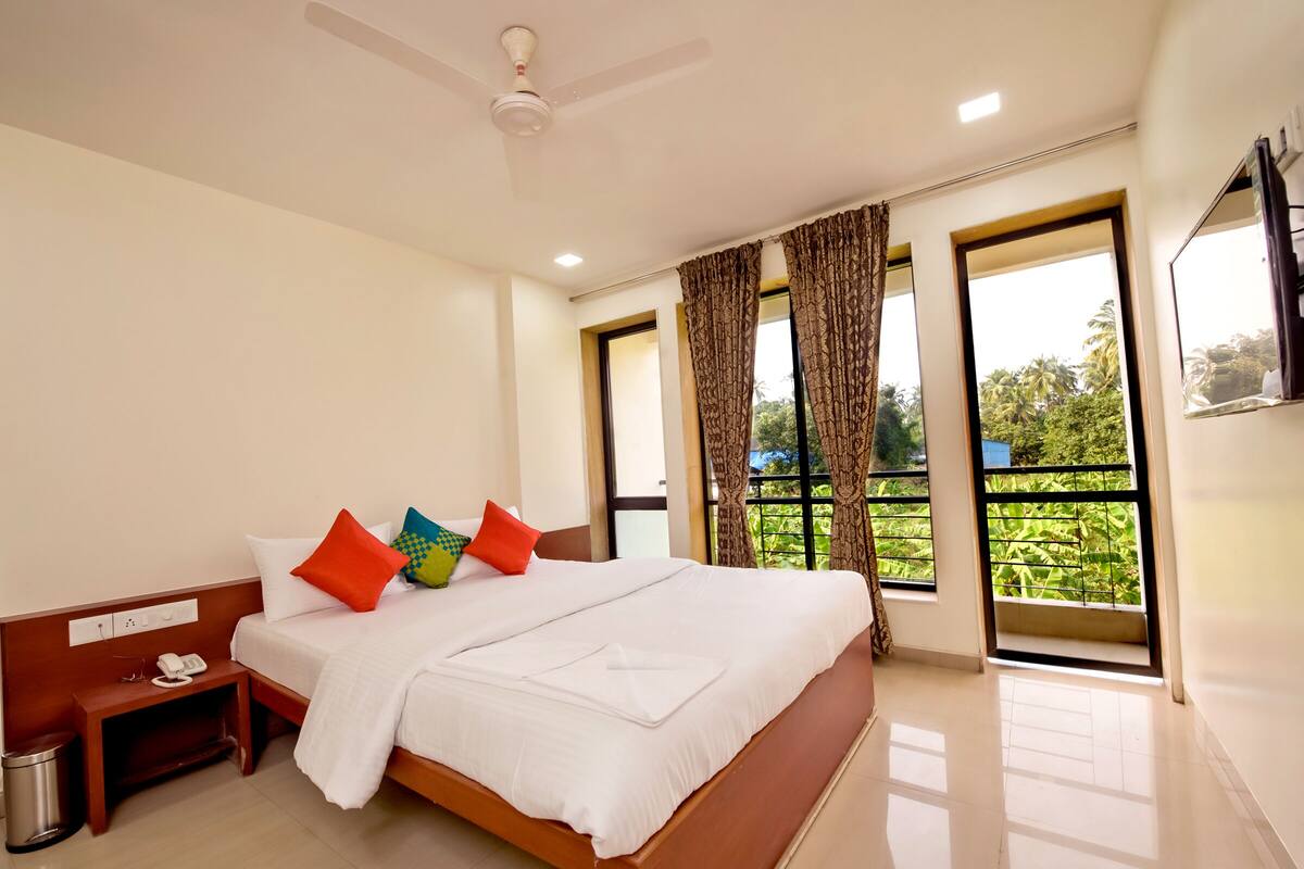Quarto de hotel com uma cama grande, uma mesa de cabeceira com telefone e vista para o verde pelas janelas.
