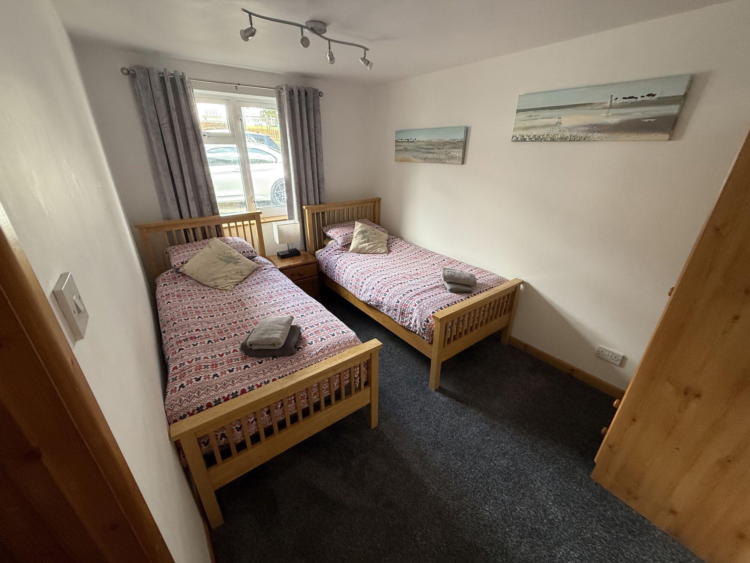 Un dormitorio con dos camas de madera, una ventana con cortinas, una lámpara de pared y dos cuadros.