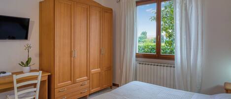 A bedroom with a bed, a wooden wardrobe, a desk with a chair, a television, and a window with curtains.