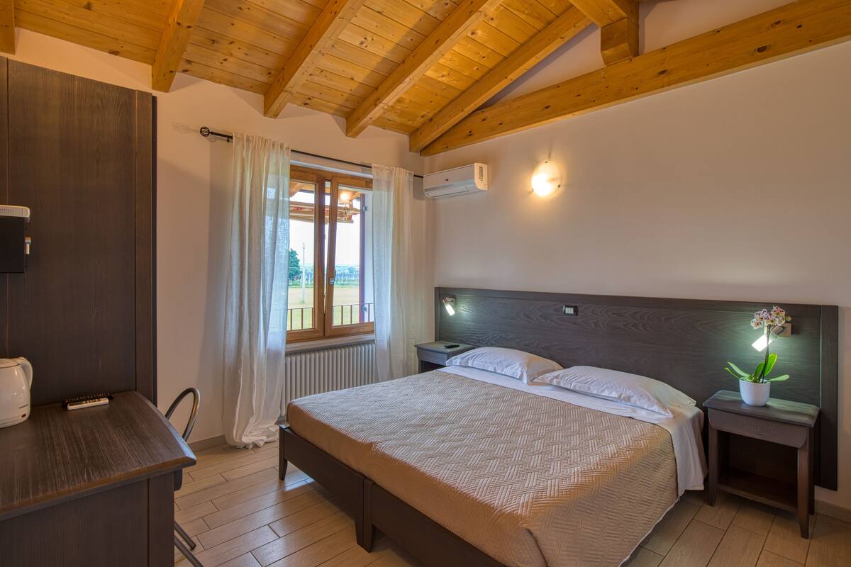 A bedroom with a wooden ceiling, a bed with white linens, a bedside table with a lamp, and a window with curtains.