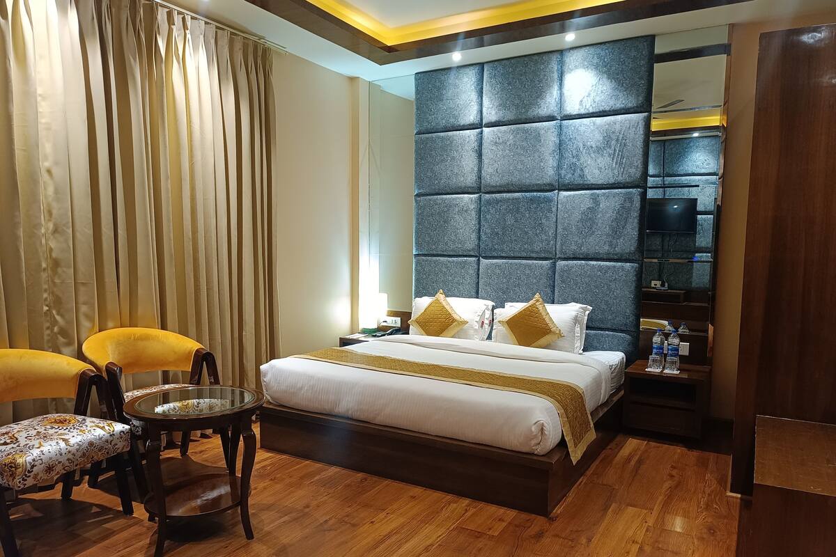 A hotel room with a large bed, a bedside table with water bottles, a chair, and a wooden floor.