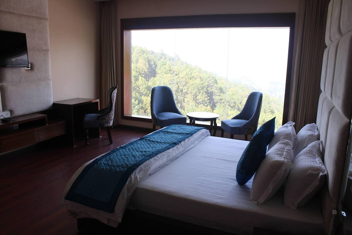 A hotel room with a large bed, two chairs, a small table, a desk, and a TV.