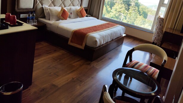 A hotel room with a large bed, a desk with a chair, a round table, and a view of trees through the window. - Mastiff Hotel Dalhousie (Dalhousie)