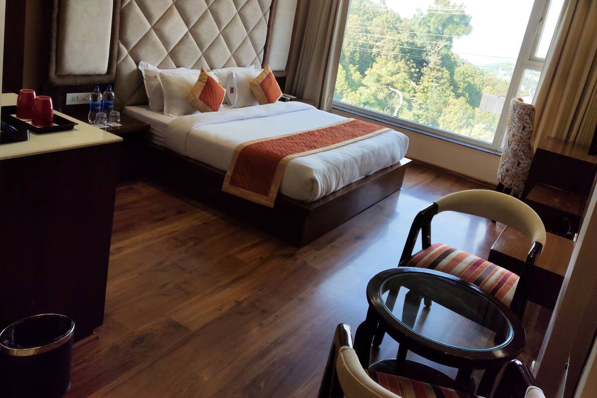 A hotel room with a large bed, a desk with a chair, a round table, and a view of trees through the window.