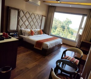 Habitación de hotel con una cama grande, un escritorio con silla, una mesa redonda y vistas a árboles por la ventana.