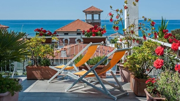 A rooftop terrace with sun loungers, potted plants, and a view of the sea and buildings. - Hotel Scandinavia (Massa)