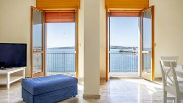 A room with a television, a blue ottoman, and a balcony with a view of the sea and a distant island.