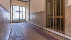 Un pasillo con piso de baldosas, un ventanal grande con barrotes de metal y una puerta de madera con paneles de vidrio.