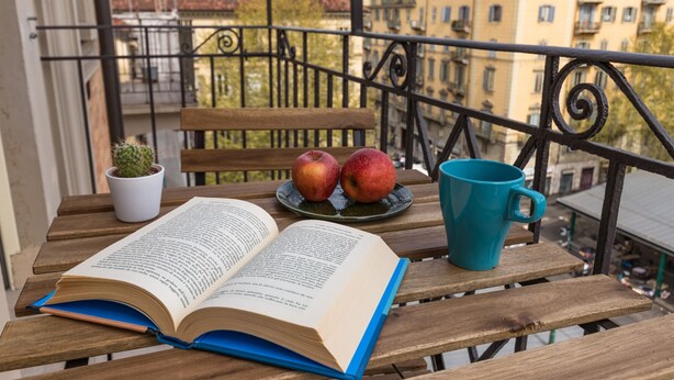 Una mesa de madera con un libro abierto, dos manzanas, una planta pequeña en maceta y una taza azul en un balcón con baranda de metal y edificios al fondo.
