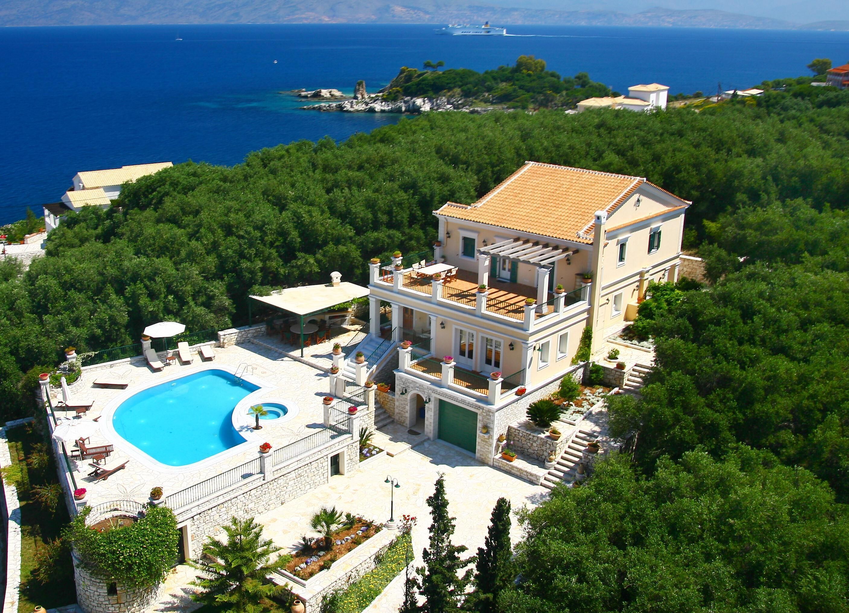 A two-story villa with a swimming pool, surrounded by lush greenery and overlooking a coastal area.