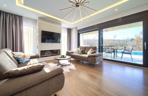 Een moderne woonkamer met een bank, een salontafel en een televisie. Er is een balkon met uitzicht op bomen.