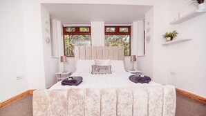 Une chambre spacieuse avec un grand lit, deux tables de chevet, une tête de lit et une vue sur la verdure par les fenêtres.