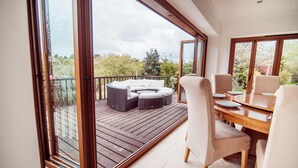 Un balcon spacieux doté d’une terrasse en bois, une table de salle à manger ronde avec des chaises, et un coin salon avec un canapé et des coussins.