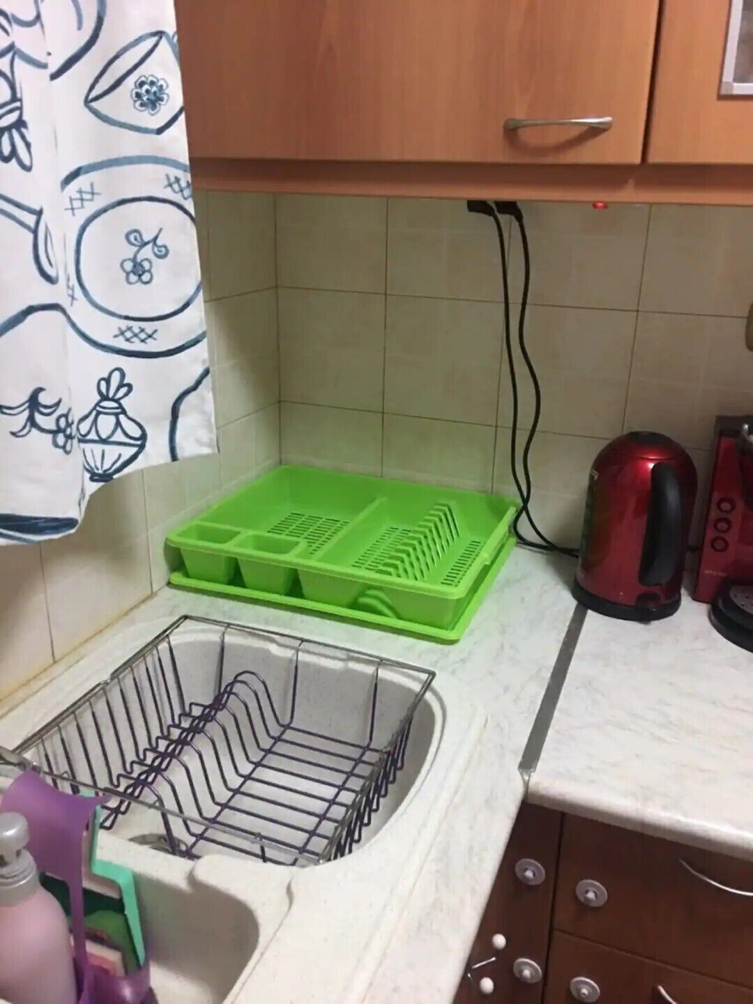 A kitchen with a sink, green plastic containers, a red kettle, and a purple bottle.