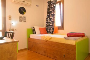 A small, single-bed room with a wooden bed frame, a desk, and a chair. There is a window with patterned curtains, a wall clock, and a wall-mounted air conditioner.
