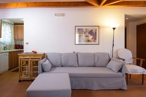 A living room with a grey sofa, a small table, a lamp, and a kitchen in the background.