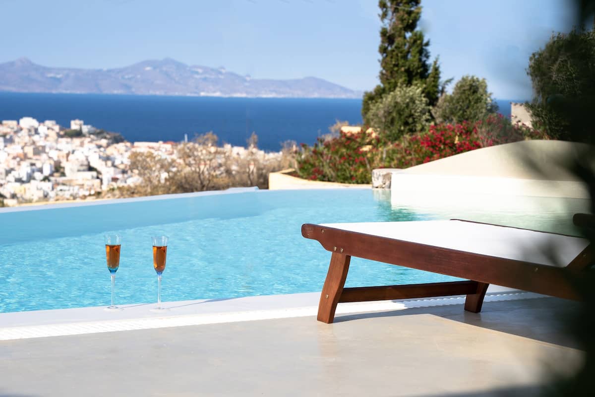 Une piscine, deux verres remplis d’une boisson, un banc en bois et une vue sur une ville côtière et des montagnes.